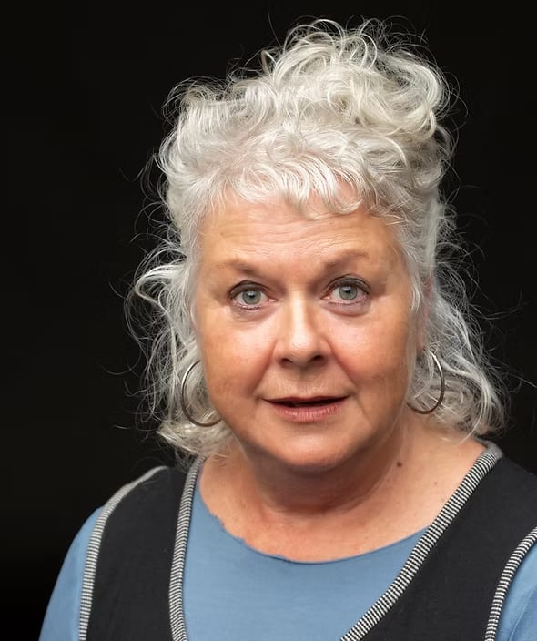 Susie Dee, a white woman with shoulder length white hair, looks at the camera. She wears a long sleeve blue top and a black dress.