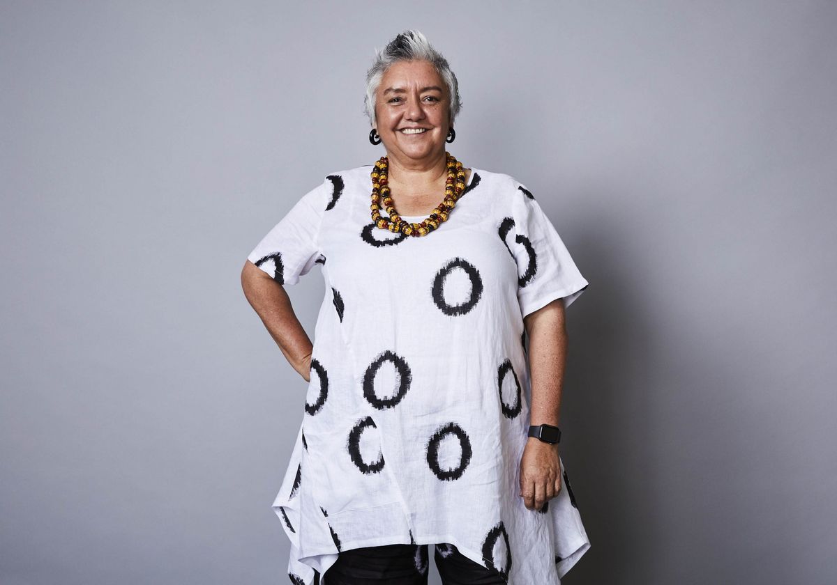 Dr Lou Bennett, an Indigenous woman with short grey hair, smiles with one hand on her hip and one resting by her side. She's wearing a white top with black circles and an orange beaded necklace and dark earrings. 