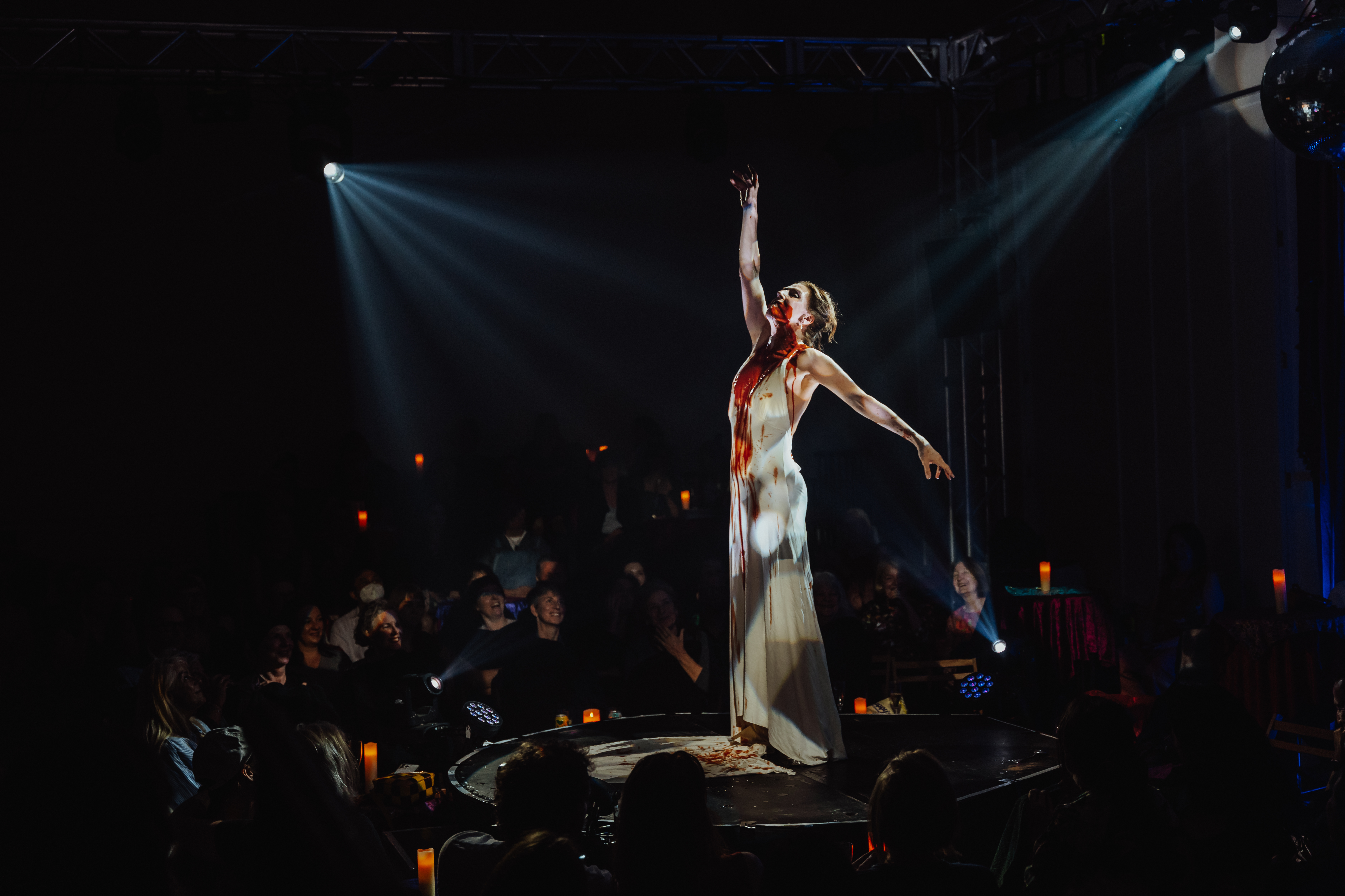 Moira Finucane, a white woman stands on a circular stage in a dark room surrounded by audience members. She wears a white dress with a red stain running down her front and stands with one arm stretched behind her and one above her.