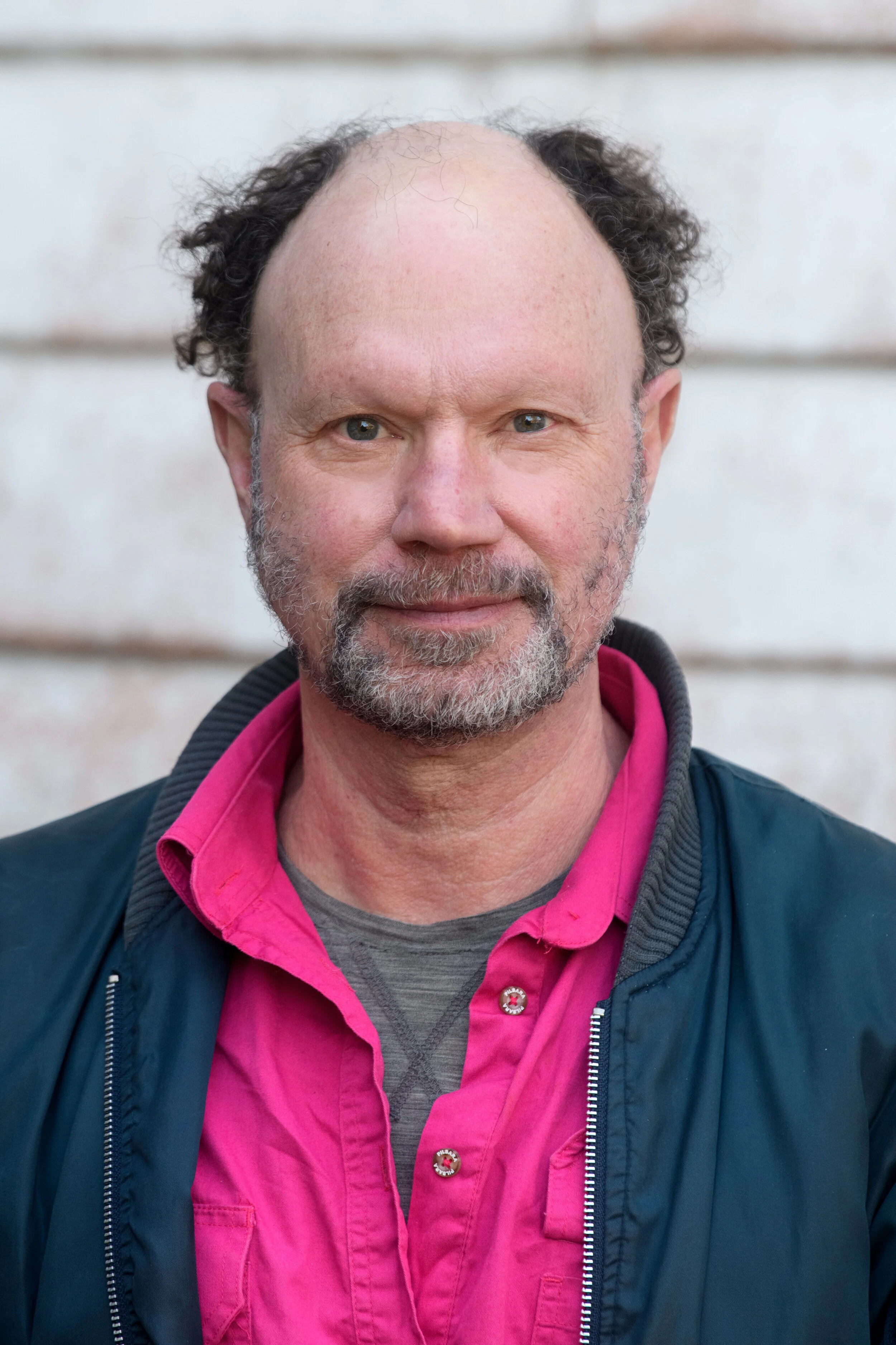 Ian Pidd, a white man with a beard, looks at the camera. He wears a pink shirt and a navy blue jacket.