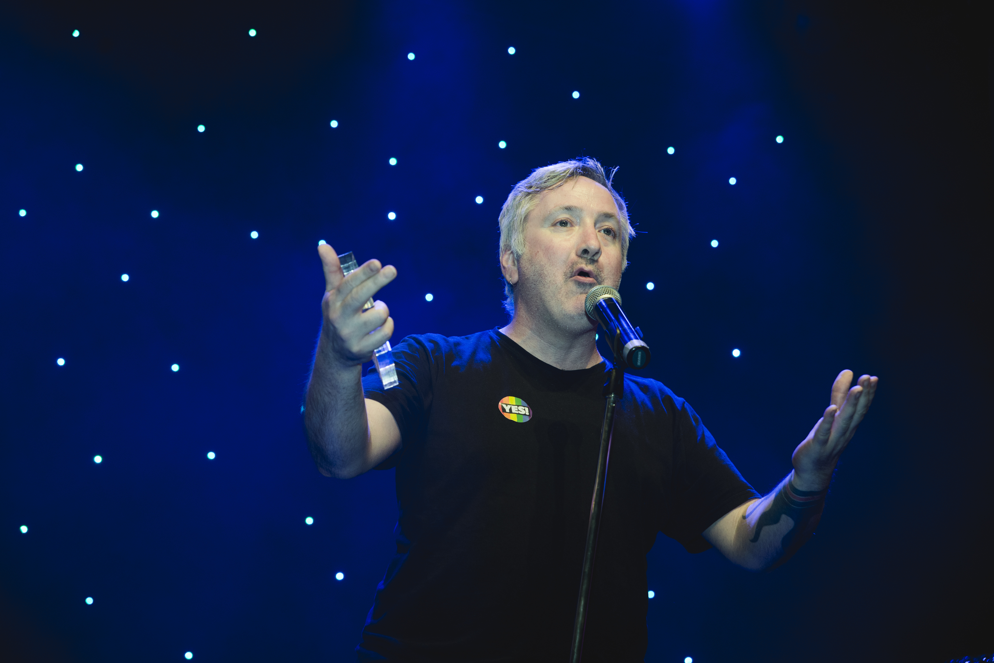 Richard Watts, a white man with short grey hair, stands on a stage with a spotlight on him. He speaks into a microphone with his hands outstretched while wearing a black t-shirt with a rainbow Vote Yes sticker on his chest.