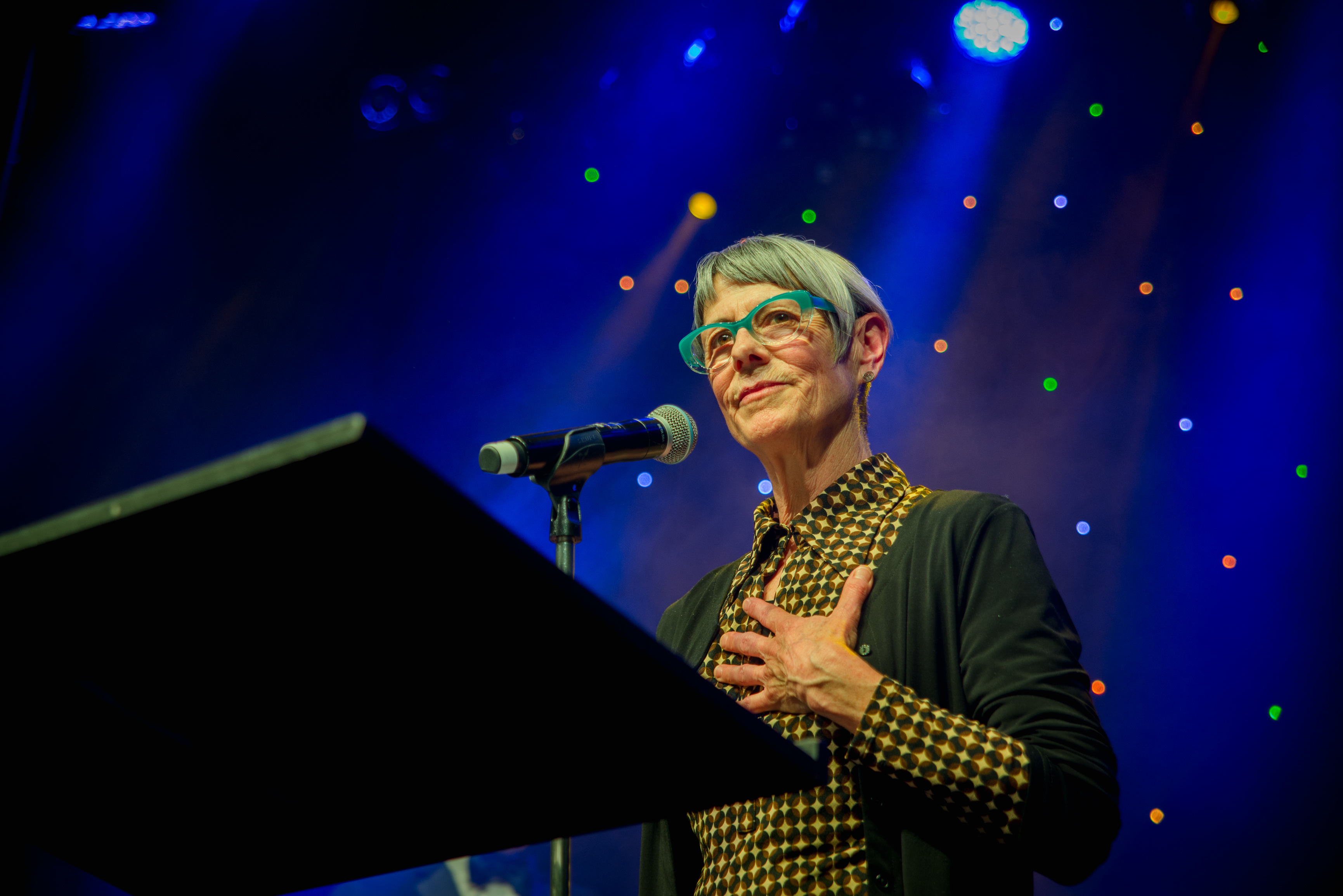 Rinske Ginsberg, a white woman with short grey hair and turquoise glasses, stands on a stage with a microphone and a lectern in front of her.